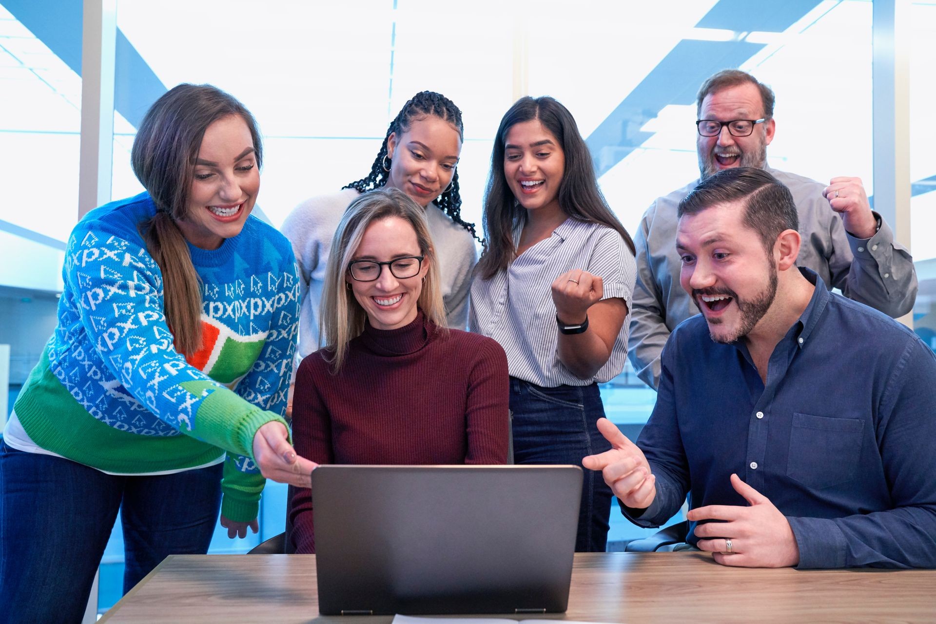 Group of excited colleagues gathered around a laptop, smiling and celebrating in a modern office setting.