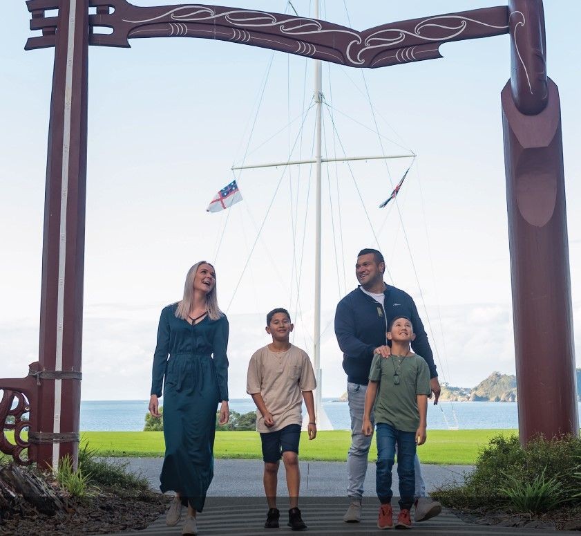 A family of four walking under a traditional wooden arch near a waterfront with flags and scenic background.