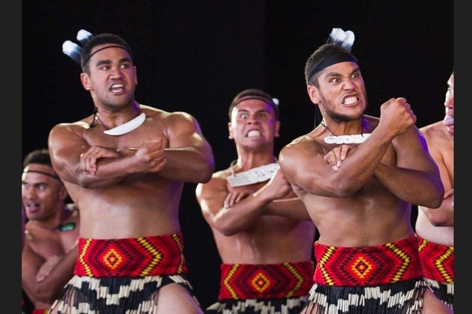 Group of men performing a traditional haka dance, wearing traditional attire with determined expressions and crossed arms.